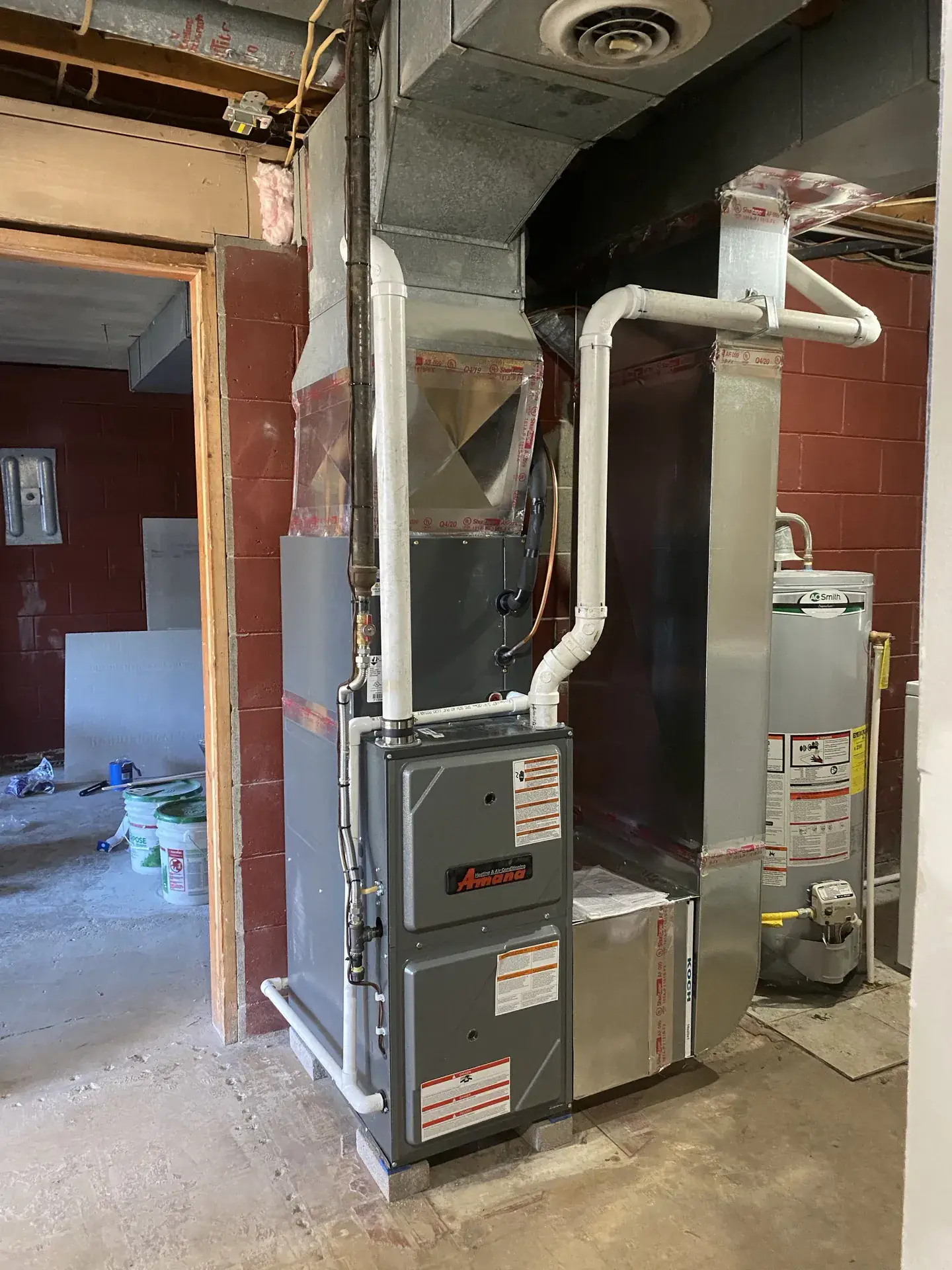 Amana high-efficiency gas furnace on a raised pad in a basement, with sealed sheet-metal plenum, media filter cabinet and white PVC intake and exhaust venting