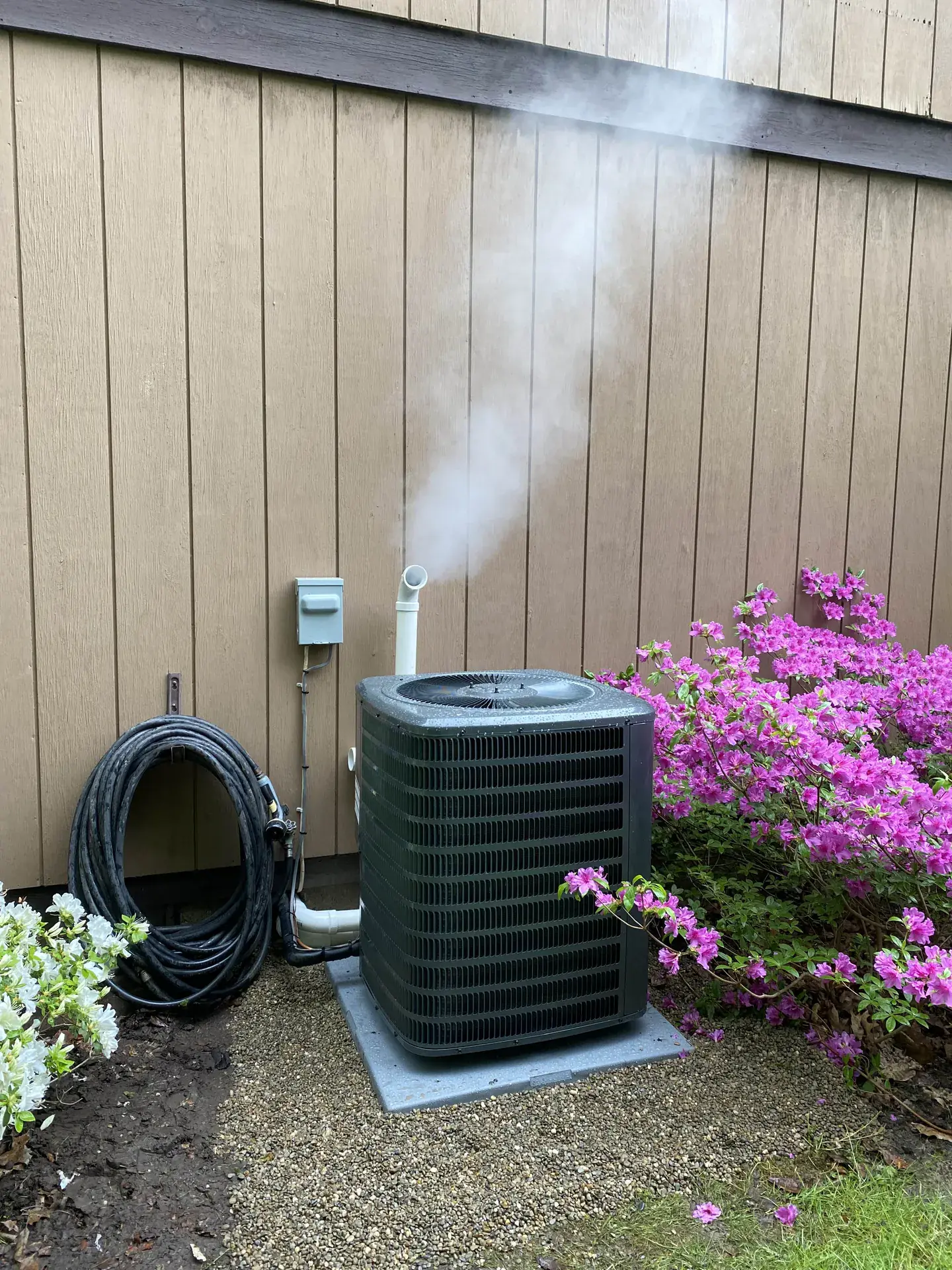 Outdoor air conditioning condenser on a composite pad beside a house with tan siding and flowering shrubs