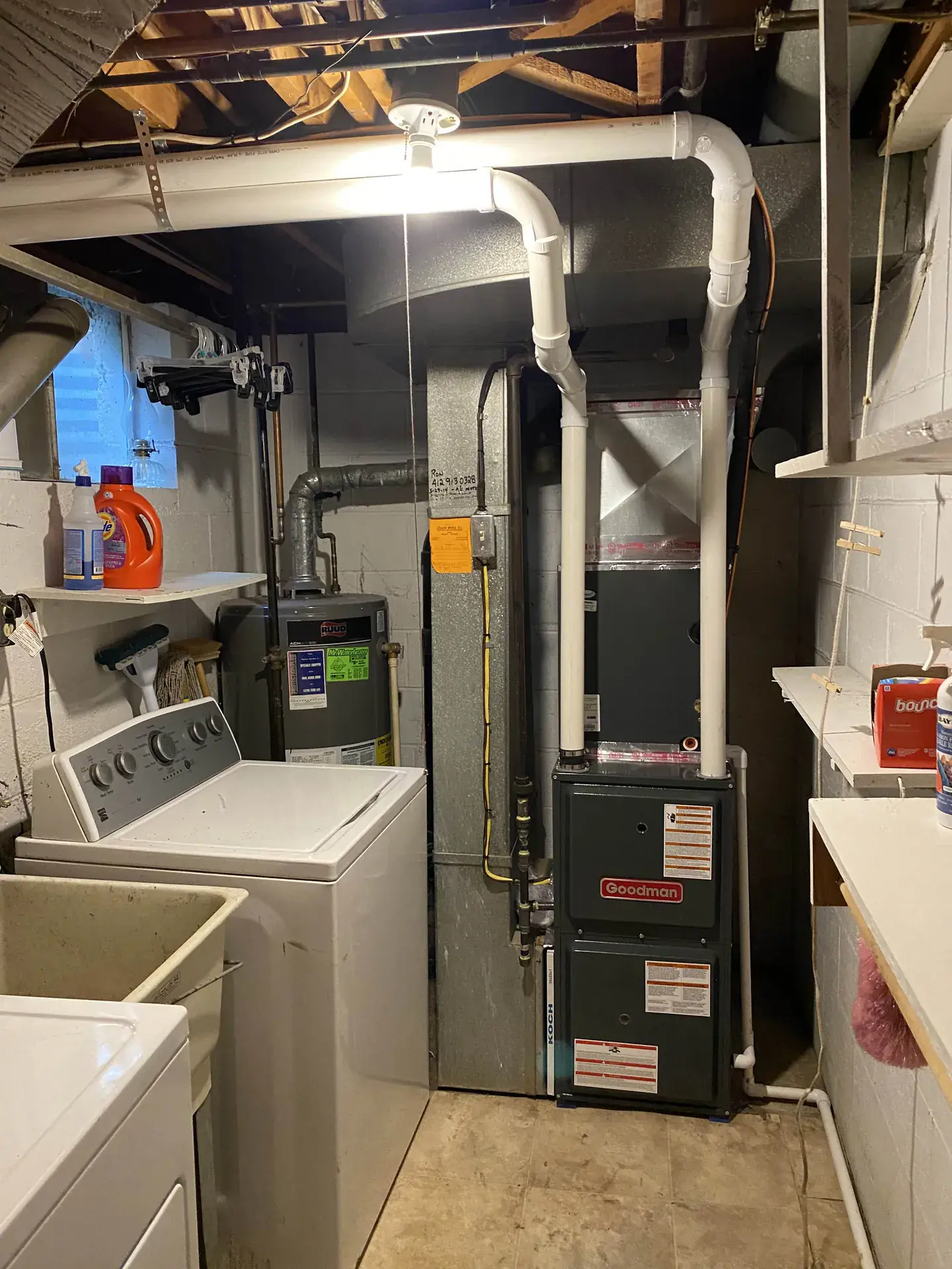 Goodman high-efficiency gas furnace installed in a narrow basement laundry room next to a water heater, with white PVC venting run along the joists