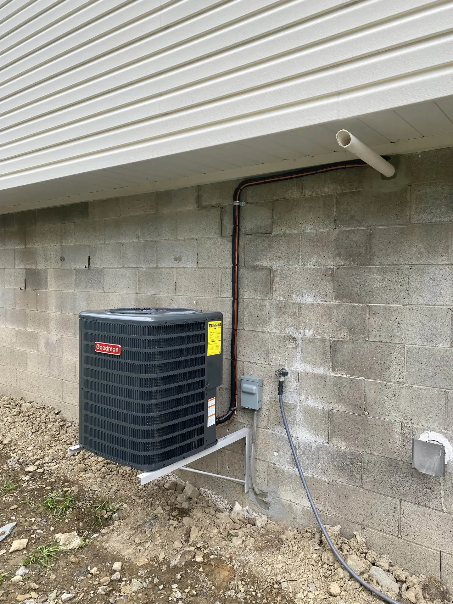 Goodman outdoor condenser mounted on a steel wall bracket against a block foundation above unfinished ground, with the line set clamped up the wall