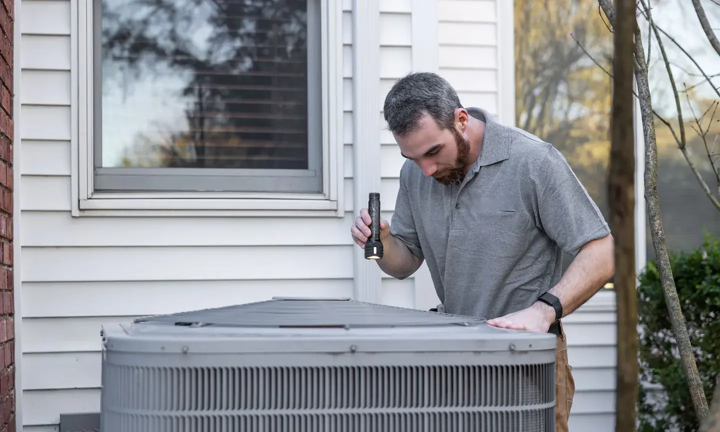 Technician inspecting the top of an outdoor air conditioning unit beside a house with a flashlight